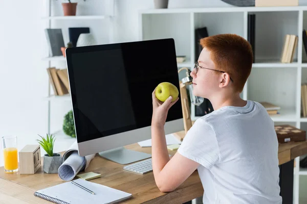 Preadolescente jengibre cabello chico celebración de manzana cerca de la computadora en casa - foto de stock