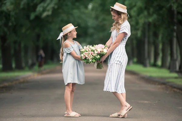 Mutter und Tochter in Strohhüten mit Blumensträußen halten Händchen im Park — Stockfoto