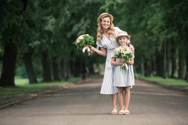 Mom and daughter in straw hats with flower bouquets posing on path in park — Stock Photo