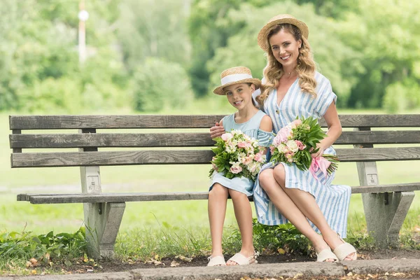 Happy stylish parent and daughter in straw hats with flower bouquets sitting on bench — Stock Photo