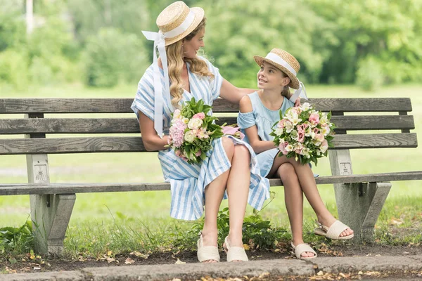 Stylish mother and daughter in straw hats with flower bouquets sitting on bench — Stock Photo