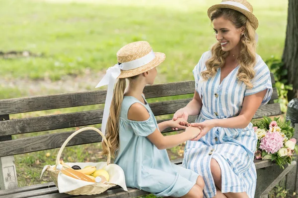 Atractiva madre y adorable hija en sombreros de paja tomados de la mano y sentados en el banco con frutas en canasta de mimbre - foto de stock