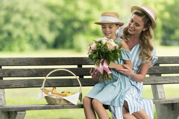 Sonrientes madre e hija con frutas en canasta de mimbre y ramo floral sentado en el banco - foto de stock