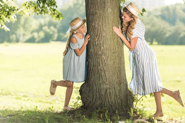 Belle mère et fille heureuse dans des chapeaux de paille posant près de l'arbre sur la pelouse verte — Photo de stock