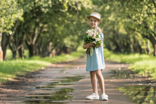 Beautiful preteen child in straw hat with bouquet of flowers posing in garden — Stock Photo