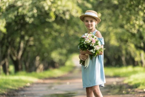 Beautiful child in dress and straw hat with flowers standing in park — Stock Photo