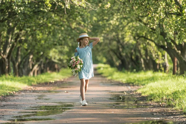 Beautiful female child in trendy dress and straw hat holding bouquet of flowers and walking in green orchard — Stock Photo