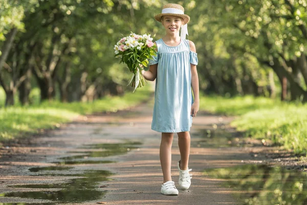 Criança pré-adolescente elegante em vestido moderno e chapéu de palha segurando buquê de flores — Fotografia de Stock