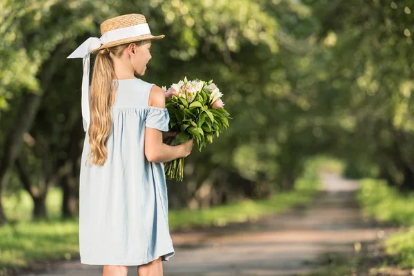 Vista trasera de niño pequeño en sombrero de paja con ramo de flores caminando en el parque - foto de stock