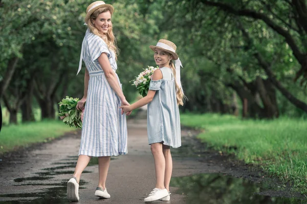 Счастливая стильная мать и дочь, держась за руки и гуляя в зеленом парке — стоковое фото