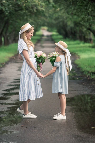 Beautiful mother and daughter in straw hats with flower bouquets holding hands and standing on path in park — Stock Photo