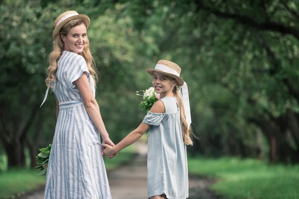 Mother and daughter in straw hats holding hands and walking in green park — Stock Photo