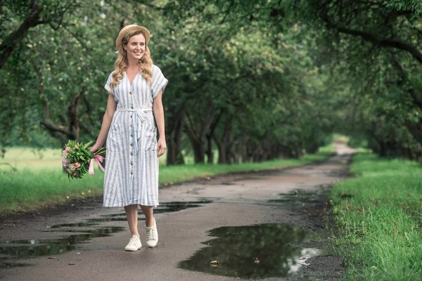 Attraktive elegante Frau im trendigen Kleid mit Blumen auf dem Weg in den Obstgarten — Stockfoto