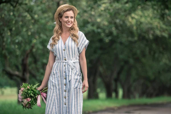 Beautiful stylish woman in dress and straw hat with bouquet standing on path in park — Stock Photo