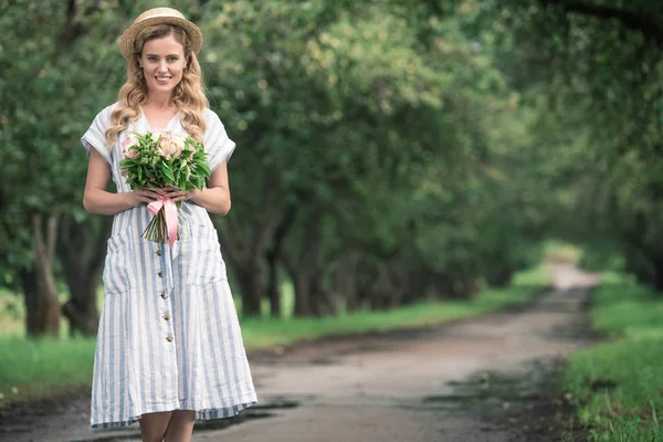 Schöne blonde Frau in Kleid und Strohhut mit Blumenstrauß, die auf dem Weg im Garten steht — Stockfoto