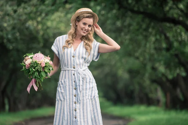 Beautiful elegant woman in straw hat with bouquet of flowers standing on path in park — Stock Photo