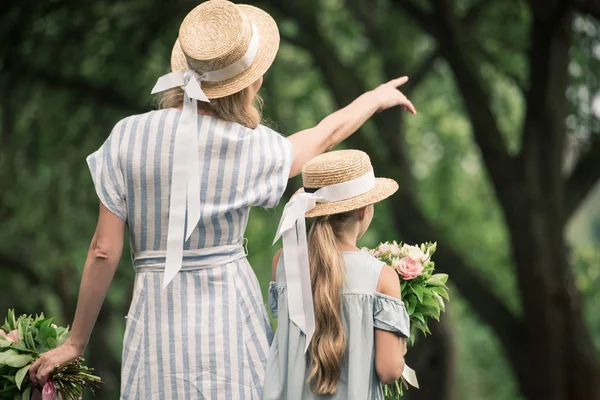 Maman et fille dans des chapeaux de paille avec des bouquets de fleurs, mère montrant quelque chose dans le parc — Photo de stock