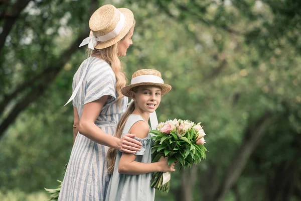 Mom and daughter in straw hats with flower bouquets walking in green park — Stock Photo