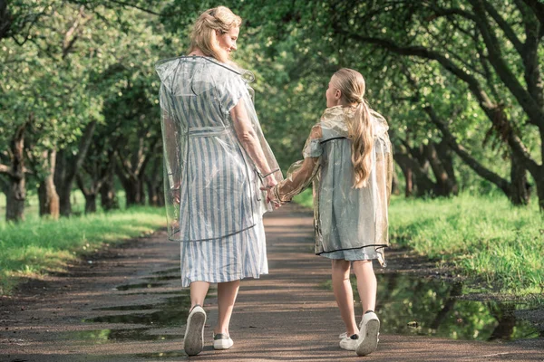 Visão traseira da mãe e da filha de mãos dadas e andando em capas transparentes na estrada molhada — Fotografia de Stock