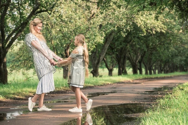 Красивая мать и очаровательная дочь в прозрачных плащах держась за руки на мокрой дороге в зеленом парке — стоковое фото