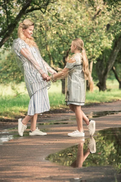 Belle mère et fille blonde en imperméables transparents tenant la main sur la route humide dans le parc — Photo de stock