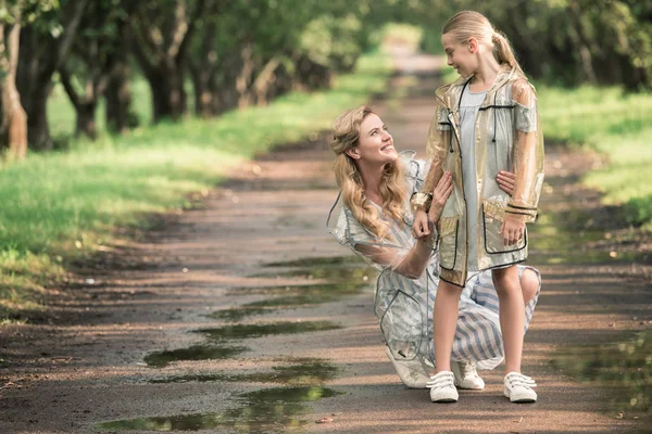 Blonde mother and adorable daughter posing in transparent raincoats on wet road in park — Stock Photo