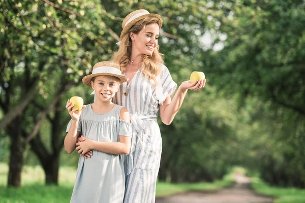 Smiling mother and daughter in straw hats looking at appples in hands — Stock Photo