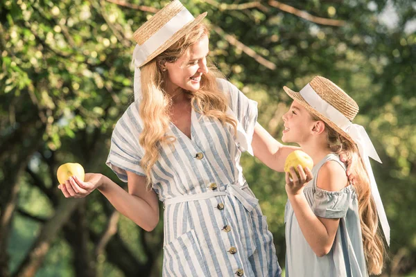 Красивая мать и дочь в соломенных шляпах держат яблоки в саду с солнечным светом — стоковое фото