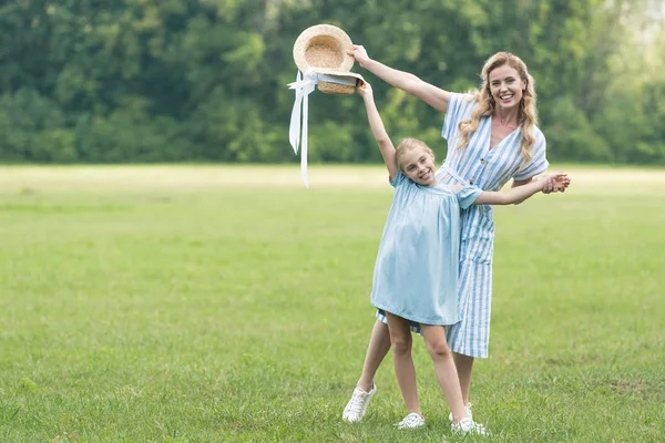 Mère heureuse et fille posant avec des chapeaux de paille sur la pelouse verte — Photo de stock