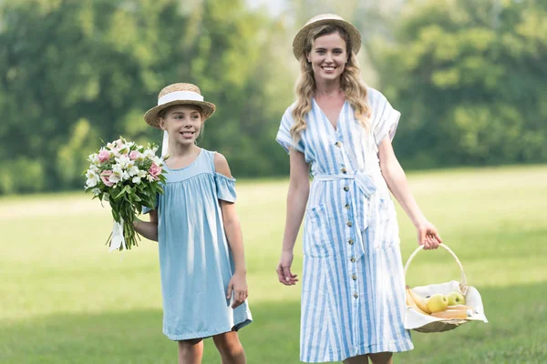 Smiling beautiful mother and daughter with fruits in wicker basket and bouquet walking on green lawn — Stock Photo
