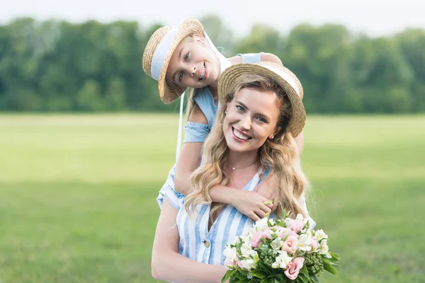 Lächelnde Mutter und Tochter posieren in Strohhüten mit Blumenstrauß — Stockfoto