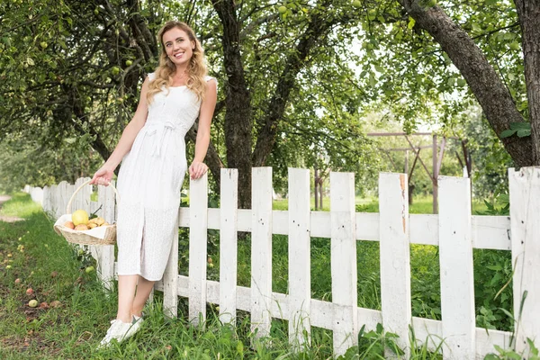 Elegante donna con frutta in vimini cesto in piedi vicino recinzione bianca in giardino — Foto stock