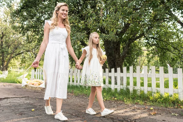 Beautiful mother and daughter with fruits in wicker basket walking near white fence — Stock Photo