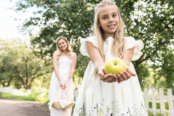 Очаровательная дочь держит яблоко, мать с плетеной корзиной на заднем плане — стоковое фото