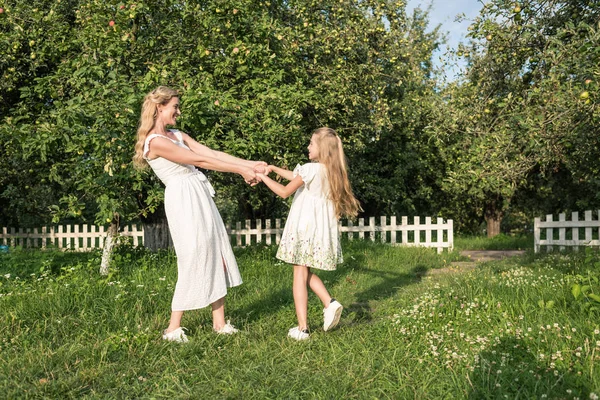 Красивая счастливая мать и дочь держатся за руки и крутятся в саду с белым забором — стоковое фото
