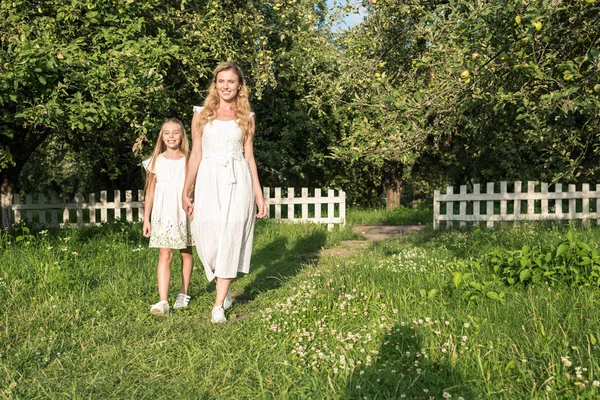 Beautiful mother and daughter in dresses holding hands in orchard with white fence — Stock Photo