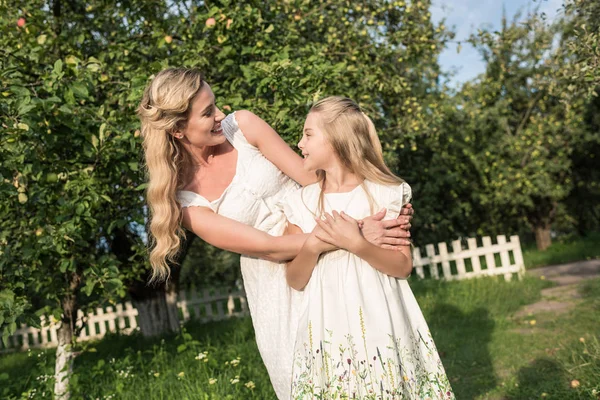 Мати і усміхнена дочка в білих сукнях, що обіймаються в сільському саду — стокове фото