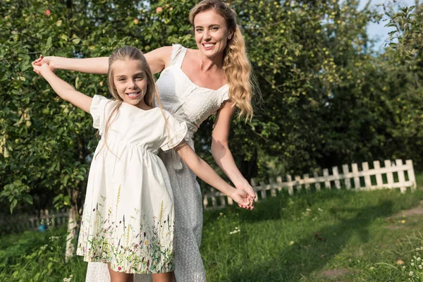 Atractiva madre y adorable hija en vestidos blancos tomados de la mano en el jardín - foto de stock