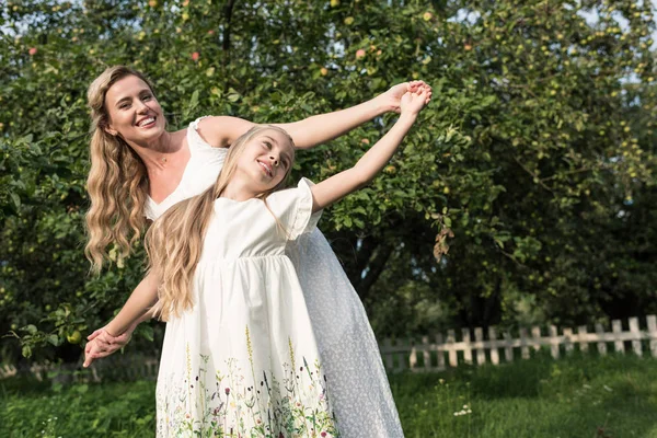 Веселая мать и дочь в белых платьях держатся за руки в саду — стоковое фото