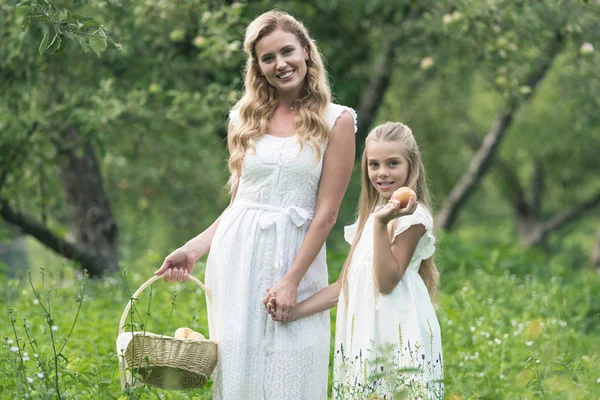 Красивая мать и дочь держат плетеную корзину с фруктами в саду — стоковое фото