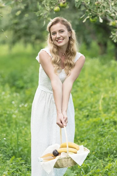 Bella donna sorridente tenendo cesto di vimini con frutta nel frutteto verde — Foto stock