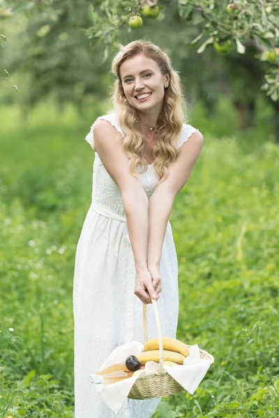 Bela mulher sorrindo segurando cesta de vime com frutas no pomar verde — Fotografia de Stock