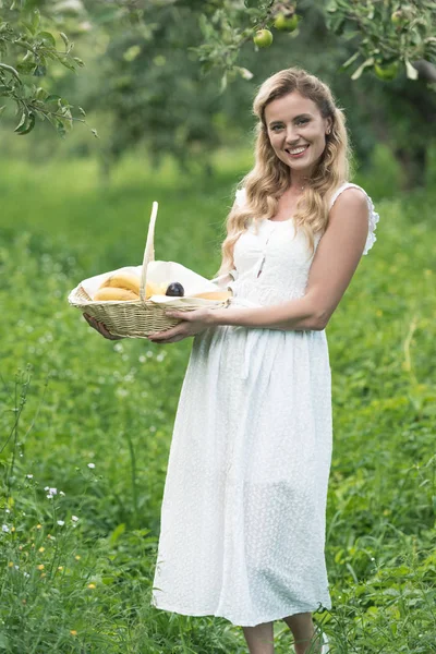 Donna felice tenendo cesto di vimini con frutta nel frutteto verde — Foto stock