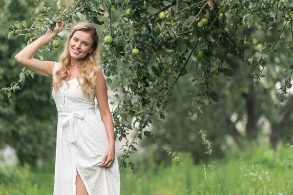Happy stylish woman in white dress posing in orchard with apple trees — Stock Photo