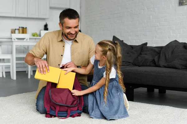 Padre sonriente ayudando a la pequeña hija a empacar la mochila para el primer día en la escuela, de vuelta al concepto escolar - foto de stock