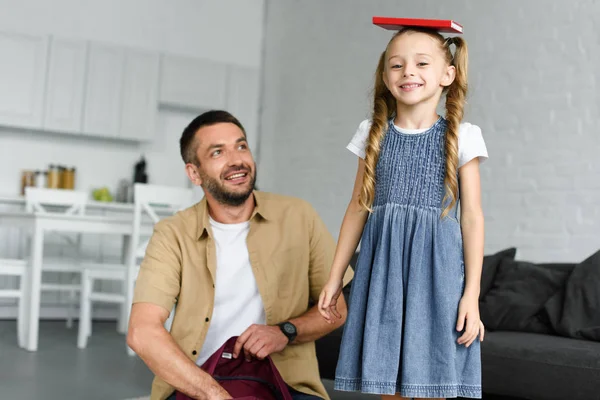 Niño sonriente con libro en la cabeza y el padre cerca en casa - foto de stock