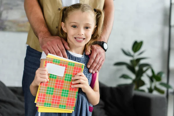 Vista parziale del padre e della figlia sorridente con i quaderni in mano a casa, ritorno al concetto di scuola — Foto stock