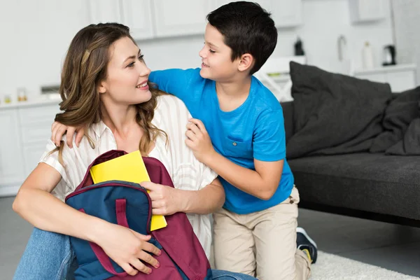 Retrato de menino sorridente abraçando a mãe com livro e mochila em casa, de volta ao conceito de escola — Fotografia de Stock