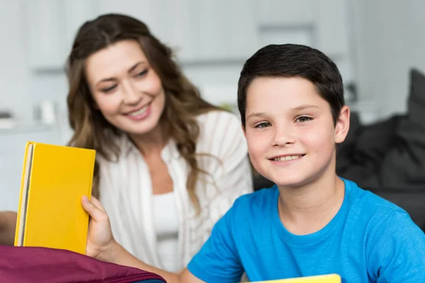 Messa a fuoco selettiva del ragazzo sorridente con libro e madre dietro a casa — Foto stock