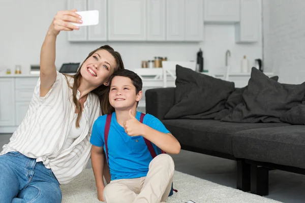 Sonriente madre e hijo con mochila y pulgar hacia arriba tomando selfie en el teléfono inteligente juntos en casa en el primer día de escuela - foto de stock